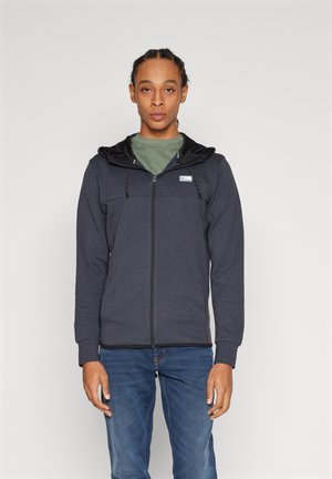 Dunkelblaue Zip-Hoodie mit einer schwarz gefütterten Kapuze, verstellbarem Kordelzug, vorderen Eingrifftaschen und gerippten Bündchen, kombiniert mit blauen Jeans.