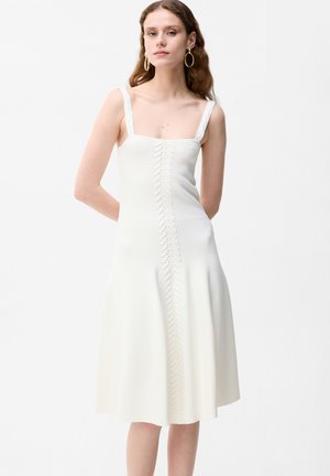 Vrouw draagt een mouwloze witte midi-jurk met brede bandjes en een verticale decoratieve detail, staand tegen een effen achtergrond.