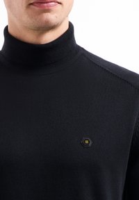 Zwarte coltrui met een gestructureerde brei, geribbelde kraag en een hexagonaal logopatch met gele accenten aan de linkerkant.