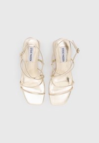 Goldene, metallische Absatzsandalen mit quadratischer Zehenform, ausgestattet mit dünnen, überkreuzten Riemen und einem Schnallenverschluss am Knöchel. Glatte Textur mit glänzendem Finish.