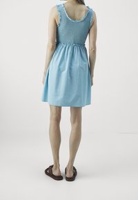 Light blue, sleeveless dress with ruffled straps and a smocked bodice, flowing into a loose skirt, made of a soft fabric. Worn with brown sandals.