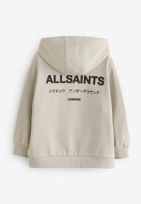 Sudadera de algodón beige con un gran logotipo negro "ALLSAINTS" en la parte posterior. Presenta una capucha, puños acanalados y cinturilla. Texto en varios idiomas.
