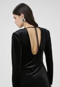 Robe en velours noir à manches longues et découpe profonde dans le dos, avec des liens fins au cou. Texture lisse avec un tombé élégant.