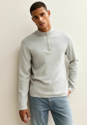 Sudadera de cremallera gris claro hecha de un tejido suave, con cuello alto y mangas largas, combinada con unos vaqueros de denimblanco claros.
