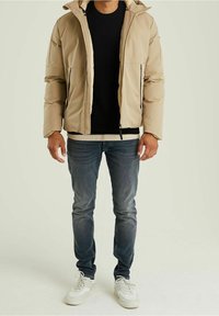 Beżowa pikowana kurtka z kapturem, zapinana na zamek, z dwoma bocznymi kieszeniami. Noszona na czarnym swetrze, w zestawie z niebieskimi dżinsami i białymi trampkami.