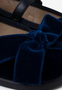 Sandalias de terciopelo azul con un gran lazo decorativo, textura suave y una correa negra con elástico. Forro interior suave en color beige y suela plana.