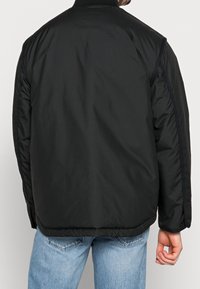 Musta tikattu takki, jossa on korkea kaulus, sileä pinta, nappiyksityiskohdat hihansuissa ja löysä istuvuus.