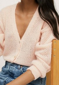 Rosa, gestrickte Strickjacke mit tiefem V-Ausschnitt, Knöpfen vorne und umgeschlagenen Ärmeln. Kombiniert mit hochtaillierten Jeansshorts.