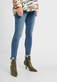 Blå tight jeans med fransade fållar, kombinerat med olivgröna ankelboots i krokodilmönster som har spetsig tå och stilettklack.