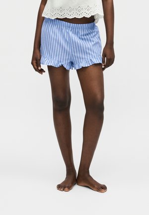 Persona con pantalones cortos de rayas verticales azules y blancas con dobladillos fruncidos y una camiseta corta blanca de encaje con ojales, descalza sobre un fondo blanco.