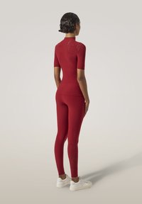 Röd åtsittande träningsset med korta ärmar och hög hals, med texturerade mönster på axlarna och leggings, i kombination med vita sneakers.