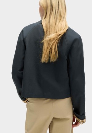 Blonde vrouw van achteren gezien die een zwart jasje met geruite manchetten en beige broek draagt tegen een effen achtergrond.