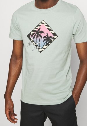Homme portant un t-shirt vert clair avec un graphique de palmier et le texte "All Day Long", associé à un pantalon noir, main dans la poche.