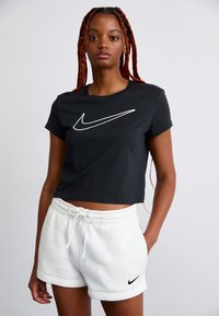 Mulher com longas tranças vermelhas, vestindo uma t-shirt preta cropped da Nike e calções brancos de cordão da Nike, a posar contra um fundo simples.