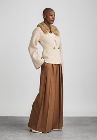 Beige double-breasted jacket with a fur collar, paired with wide brown trousers. The fabric appears soft and textured with a tailored fit.