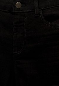 Gros plan sur un jean en denim noir montrant la ceinture, le bouton avec le nom de la marque, la passants de ceinture et les détails cousus de la poche avant.