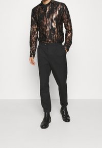 Camisa negra con estampado y acentos en bronce metálico, combinada con pantalones oscuros ajustados y botas negras de tobillo. Diseño ajustado, textura suave.