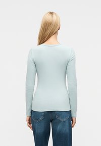 Γυναίκα με μακριά ξανθά μαλλιά, φοράει εφαρμοστή μακρυμάνικη μπλούζα ανοιχτού γαλάζιου χρώματος και μπλε τζιν, στέκεται γυρισμένη πλάτη σε απλό φόντο.