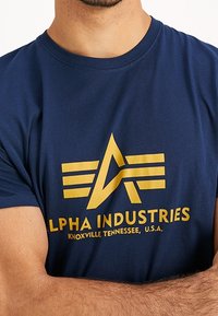 Námořnická modrá bavlněná tričko s žlutým grafickým logem, které obsahuje stylizované "A" a pruhy. Text zahrnuje "ALPHA INDUSTRIES, KNOXVILLE, TENNESSEE, U.S.A."