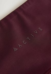 Bordo platneni džep s utisnutim tekstom "ACTIVE"; glatka tekstura s suptilnim sjajem, pojačano šivanje i trokutasti naglasak.