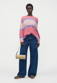 Femme portant un pull rayé rose et bleu, un jean bleu à jambes larges, des baskets vertes, et tenant un sac à main cylindrique beige.