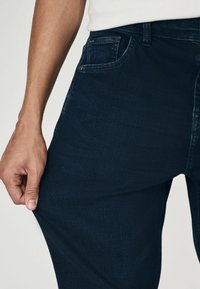 Jeans din denim albastru marin cu un design ajustat. Caracteristici un buzunar frontal și o ușoară elasticitate, arătând o mână care trage de țesătură pentru a demonstra flexibilitatea.