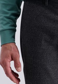 Main droite d'une personne portant une chemise à manches longues verte reposant à côté de pantalons texturés sombres.