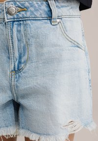 Hellblaue Denim-Shorts mit fransigem Saum, Fünf-Taschen-Design und silberner Knopfverschluss. Verfügt über gelbe Nähte.