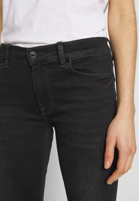 Gros plan sur une personne portant un jean en denim noir et une chemise blanche, montrant la taille, le bouton et la main droite reposant sur le côté.