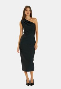 Vestido midi negro de un solo hombro con una silueta ajustada, textura suave y detalles drapeados en el hombro. Combinado con zapatos de tacón alto.