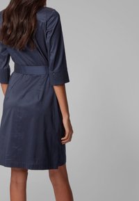 Granatowa sukienka z rękawami o długości trzech ćwiartek, z paskiem w talii i gładkim, teksturowanym materiałem. Widok z tyłu ukazuje dopasowany krój i szczegóły szwów.