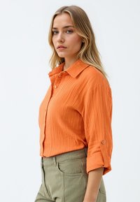Oranges Hemd mit strukturiertem Stoff, hochgeschobenen Ärmeln und einem Spread-Kragen, kombiniert mit olivgrünen Hosen.