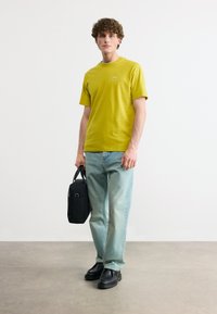 Νεαρός άνδρας όρθιος, φοράει κίτρινη μπλούζα, ανοιχτό μπλε τζιν παντελόνι, μαύρα παπούτσια και κρατάει μια μαύρη τσάντα σε απλό λευκό φόντο.