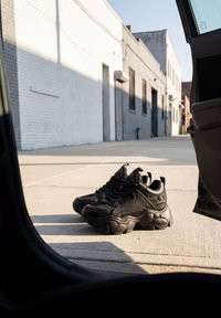 Par sorte chunky sneakers placeret på en solbelyst betonfortov ved siden af en åben bildør i en urban gyde.