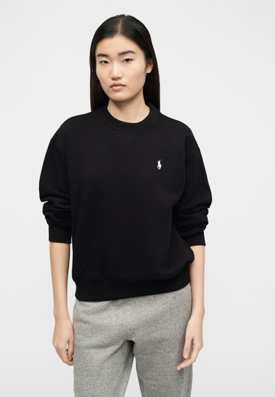 Γυναίκα με μακριά σκούρα μαλλιά φοράει μαύρο φούτερ με λαιμόκοψη και μικρό λευκό λογότυπο, καθώς και γκρι φόρμα, μπροστά σε απλό ανοιχτό φόντο.