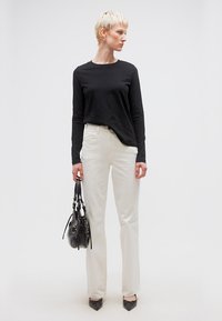 Άτομο με κοντά ξανθά μαλλιά, φορώντας μαύρο πουκάμισο με μακριά μανίκια, λευκό παντελόνι, μαύρα μυτερά παπούτσια, κρατώντας μαύρη τσάντα, στέκεται μπροστά σε απλό φόντο.