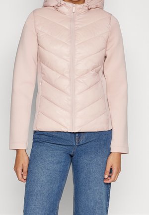 Femme portant une veste matelassée à capuche rose clair avec des coutures diagonales et un jean bleu, debout devant un fond uni.