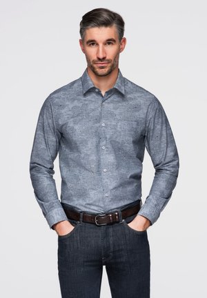 Hombre con cabello y barba gris, vistiendo una camisa azul ajustada de manga larga con estampado, metida dentro de unos vaqueros oscuros con un cinturón marrón, con las manos en los bolsillos.