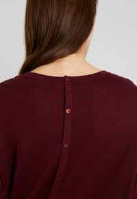 Burgundin neulepusero, jossa on pyöreä kaula-aukko, takana viisi nappikoristetta ja pehmeä, teksturoitu kangas.