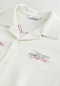 Chemise écrue à col avec boutons, avec un texte brodé en noir et rouge indiquant "ATELIER No.12 NOUVEAU EDITION Original."