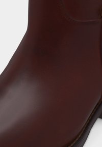 Κοντινό πλάνο της μύτης ενός λείου, σκούρου καφέ δερμάτινου μποτάκι με ορατή ραφή κατά μήκος της άκρης της σόλας σε λευκό φόντο.