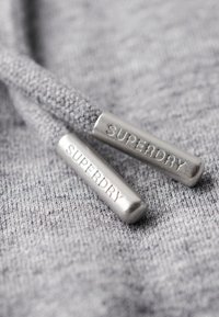 Tessuto grigio con texture a maglia che presenta due terminali metallici con la scritta "SUPERDRY" impressa, attaccati a cordini coordinati.