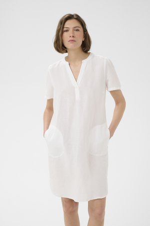 Femme debout, mains dans les poches, portant une robe blanche ample à manches courtes, sans col, avec un décolleté en V et deux grandes poches avant.