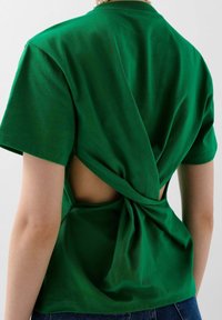 Zielona bawełniana koszulka z twisted back design, otwartą sekcją, krótkimi rękawami i okrągłym dekoltem. Widoczna para z dżinsami.