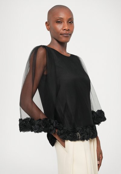 Black blouse with sheer tulle sleeves and floral embellishments at the hem. Smooth fabric, round neckline, and details feature textured flowers.