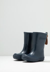 Botas de lluvia de goma azul marino con una textura lisa, de altura hasta el tobillo y suela resistente; cuenta con un borde texturizado y una etiqueta de cuero marrón.