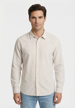 Hombre que lleva una camisa de manga larga con botones y rayas verticales beige, azul y naranja, combinada con vaqueros azul claro, posando contra un fondo blanco liso.