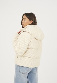 Veste matelassée en velours côtelé crème avec des côtes verticales, une grande capuche et des poches latérales, associée à un jean en denim bleu clair.