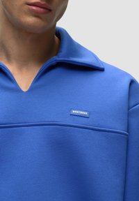 Felpa blu con colletto, dotata di una grande tasca sul petto e un'etichetta con il logo "WRSTBHVR". Texture liscia, design casual.