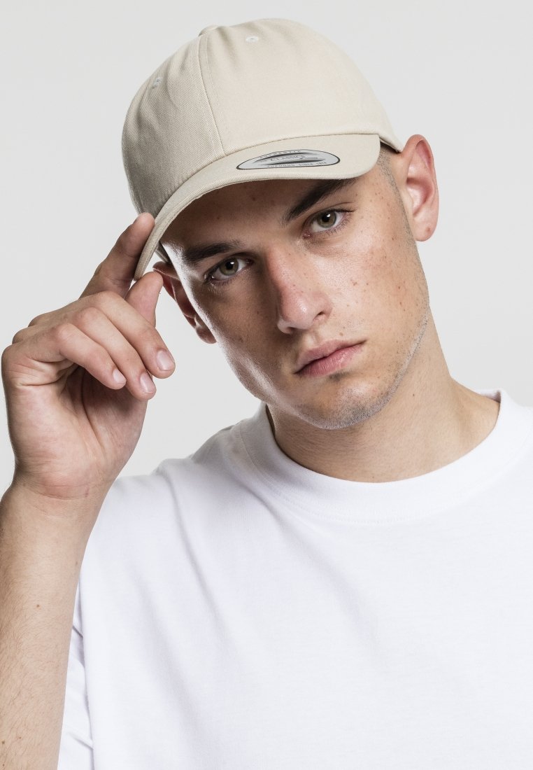 Beige baseball cap with a curved visor and a light fabric, worn by a person in a white t-shirt, hand raised to the cap's brim.